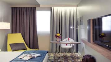 Habitación de hotel con cama, una silla, una mesita con un florero, televisión y maleta.
