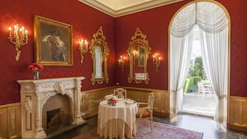 A room with a fireplace, a dining table with chairs, a large framed portrait, and ornate wall sconces.