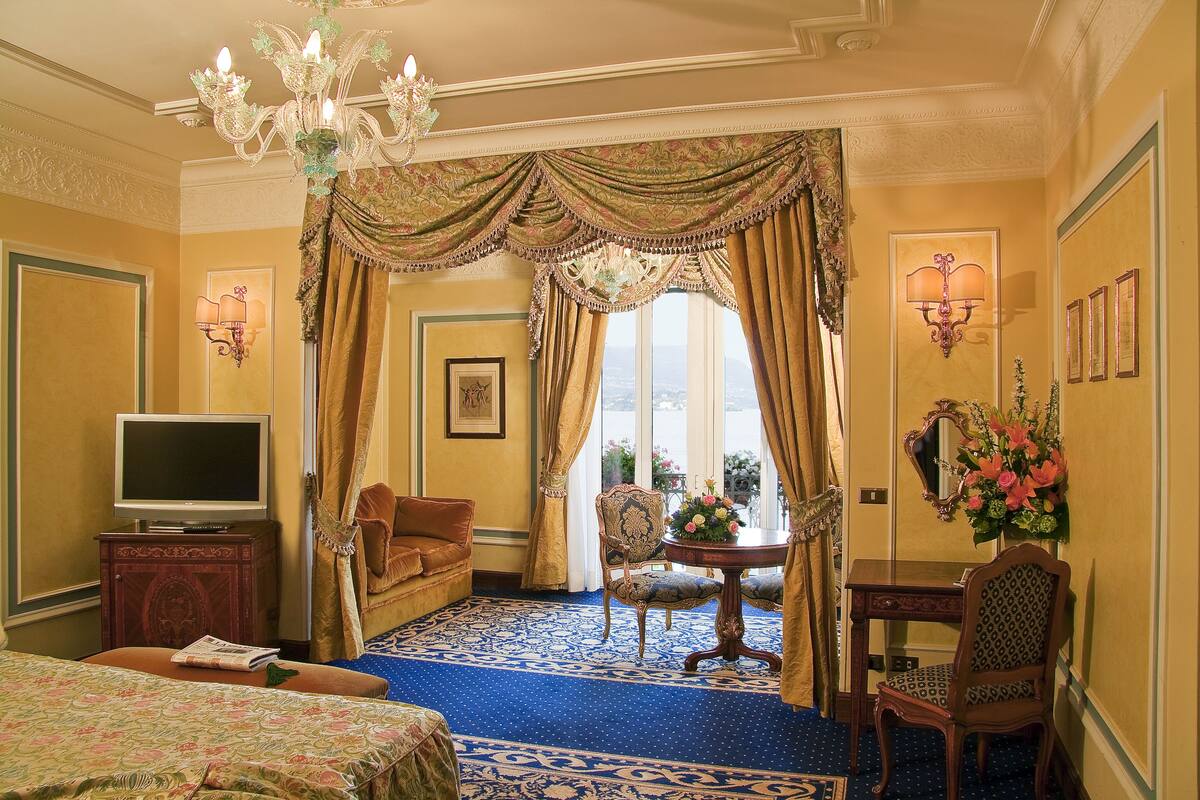 A hotel room with a large bed, a television on a wooden cabinet, a desk with a chair, and a view of the outdoors through a large window.
