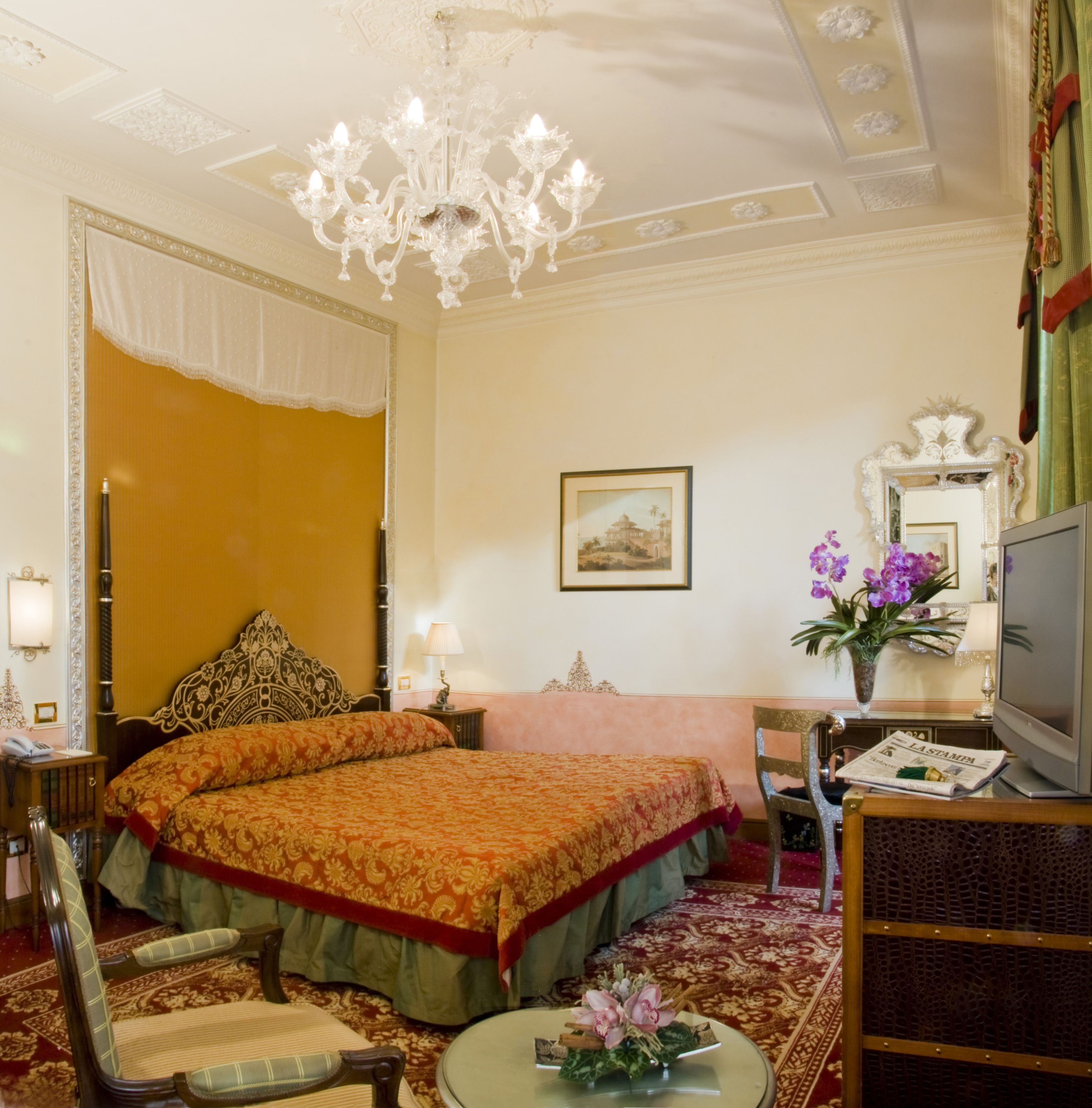 A hotel room with a large bed, a chandelier, a television, and a framed picture on the wall.