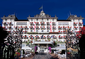 A grand hotel with multiple balconies adorned with red flowers, a central entrance with a decorative canopy, and outdoor seating areas. - Grand Hotel Des Iles Borromees (Stresa)
