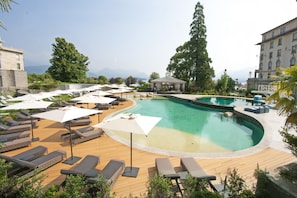 Relax by the pool surrounded by lounge chairs and umbrellas, with a grand building and lush greenery in the background.