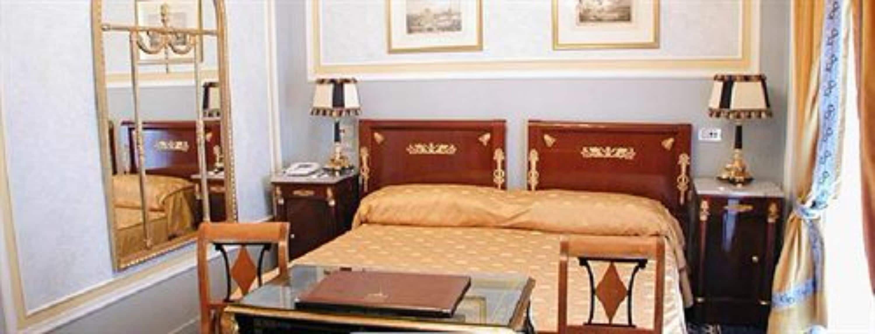 A hotel room with a double bed, a desk with a laptop, a chair, a nightstand with a lamp, and framed pictures on the wall.