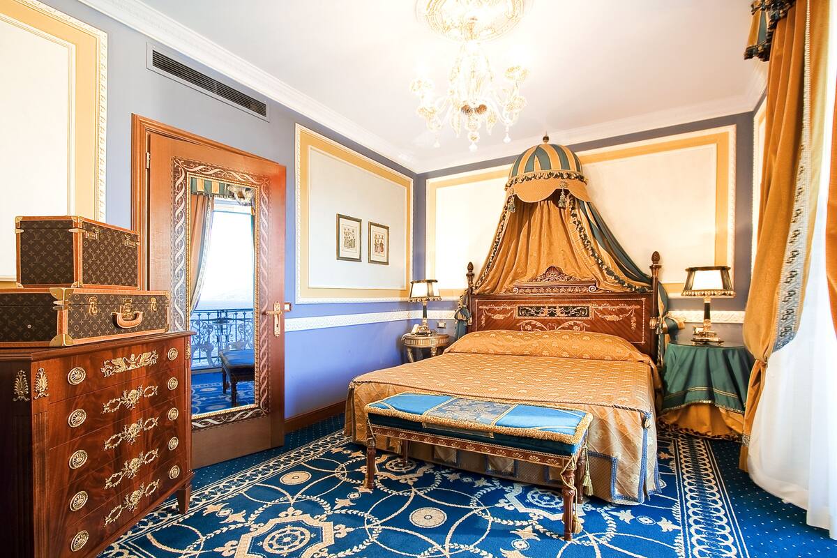 A room with a four-poster bed, a wooden dresser, a chandelier, and a patterned carpet.