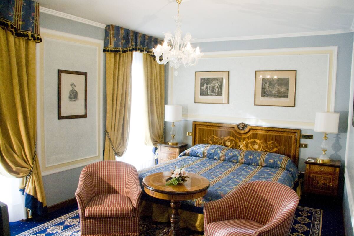 A hotel room with a large bed, two chairs, a round table, and a chandelier.