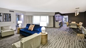 A hotel room with a blue sofa, a beige armchair, a small round table with a lamp, a conference table with chairs, and a kitchenette in the background. - Embassy Suites Hotel Charlotte (Charlotte)