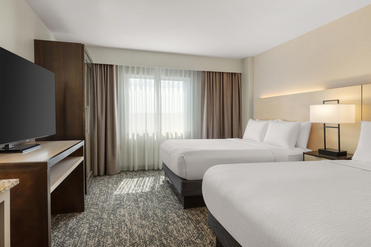 A hotel room with a flat-screen TV on a wooden desk, two beds with white linens, a small bedside table with a lamp, and a large window with sheer curtains.