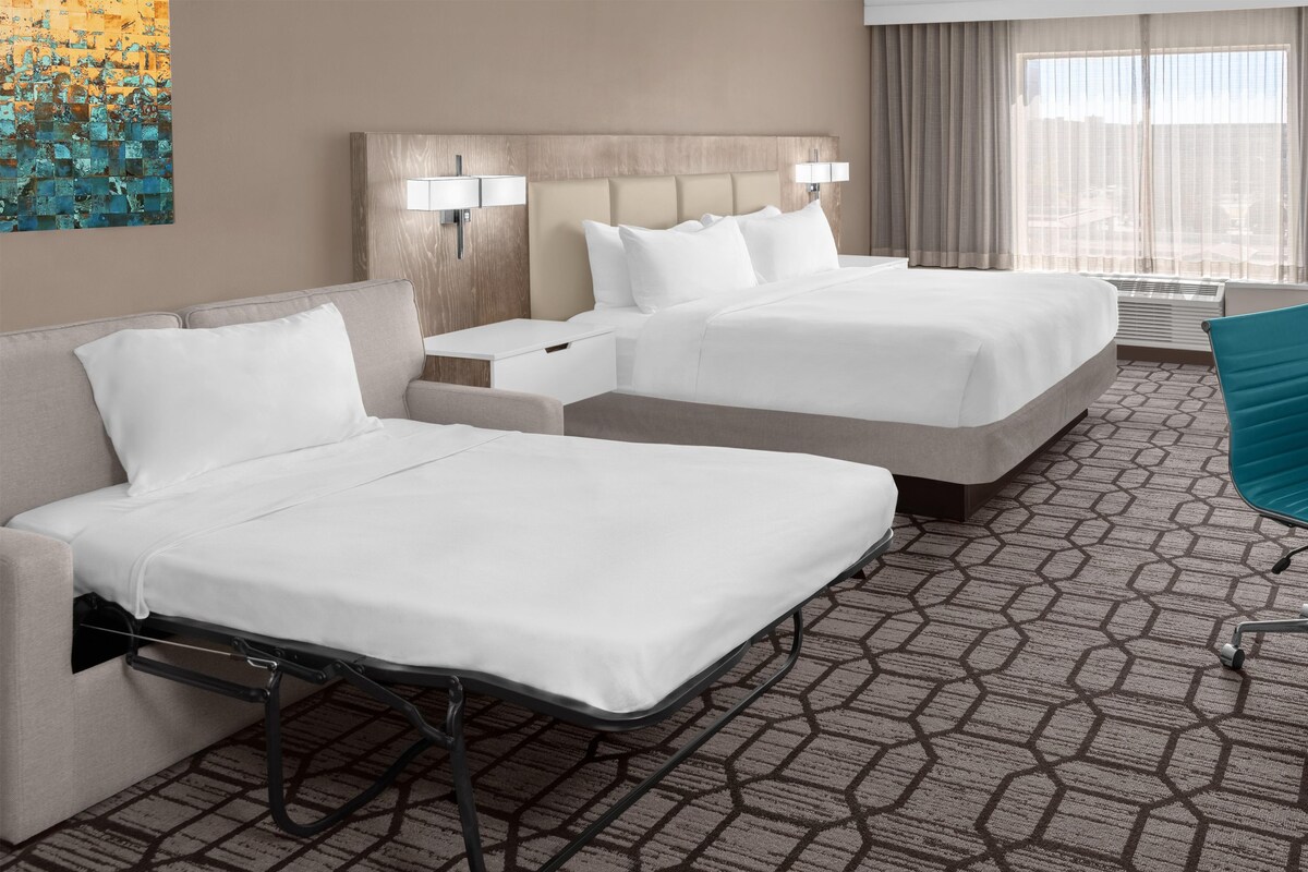 A hotel room with two beds, a desk, a chair, and a large window with curtains.