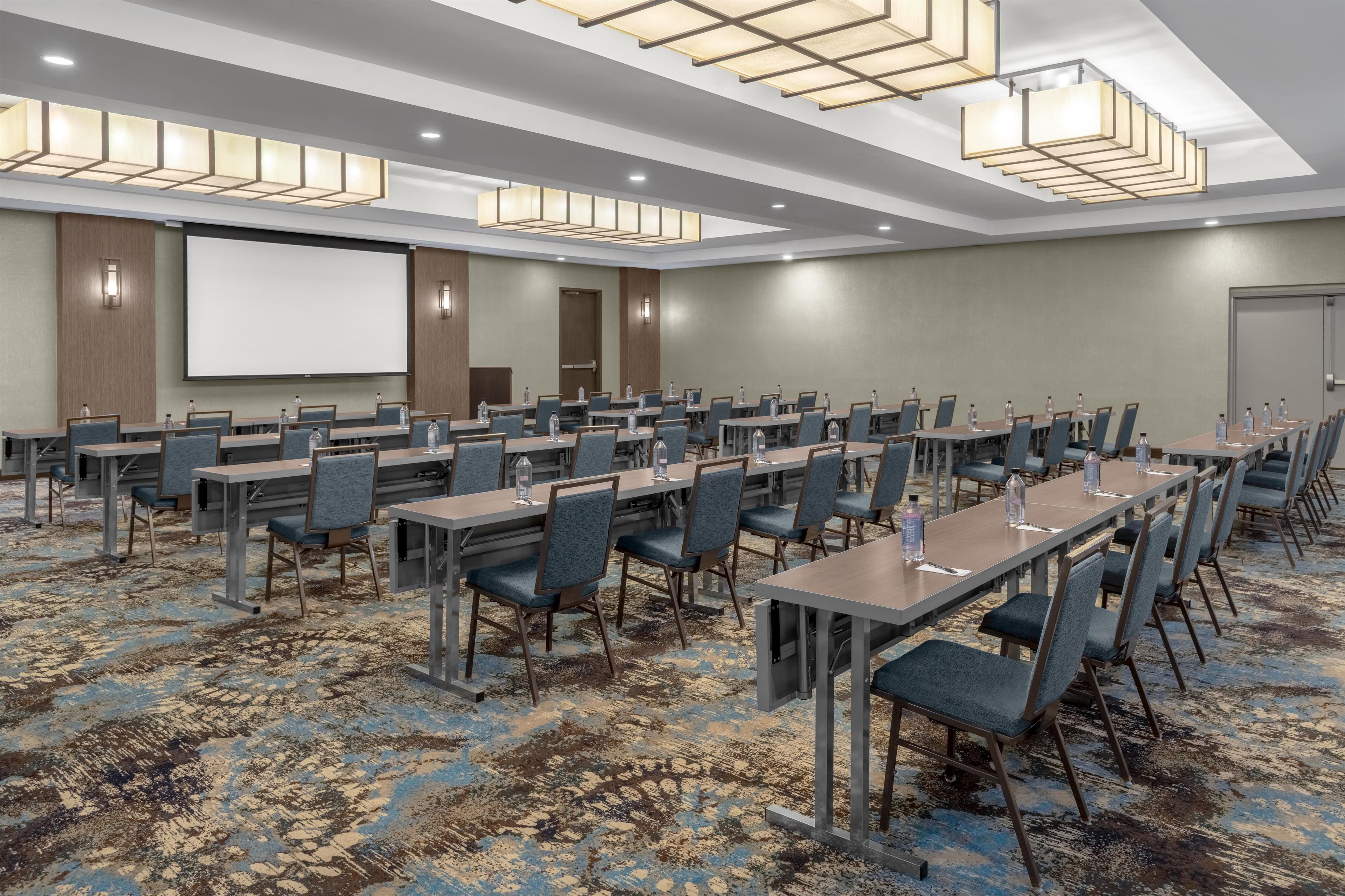 A conference room with multiple tables and chairs, a projector screen, and a carpeted floor.