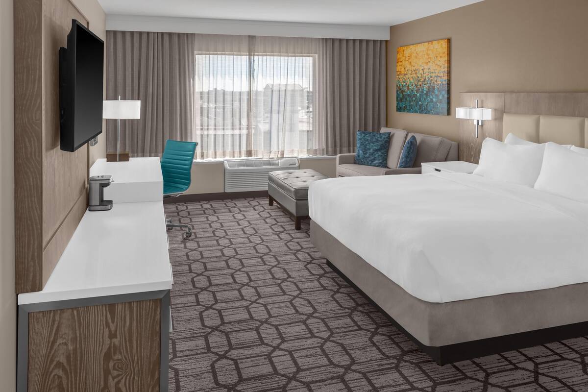 A hotel room with a large bed, a desk, a chair, a TV, a window with curtains, and a painting on the wall.