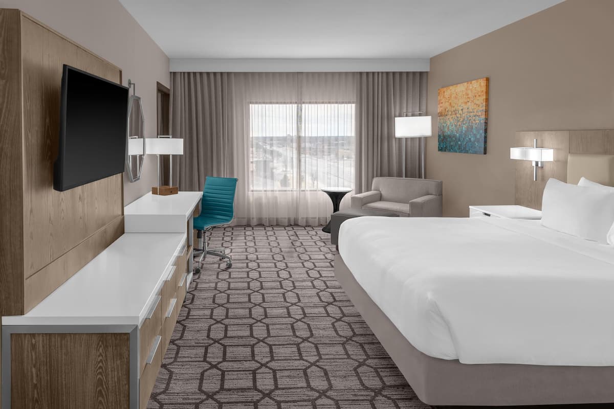 A hotel room with a bed, a desk, a chair, a TV, and a painting on the wall.