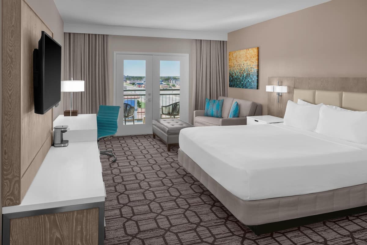 A hotel room with a large bed, a desk with a lamp, a TV mounted on the wall, a balcony with a view, and a painting on the wall.