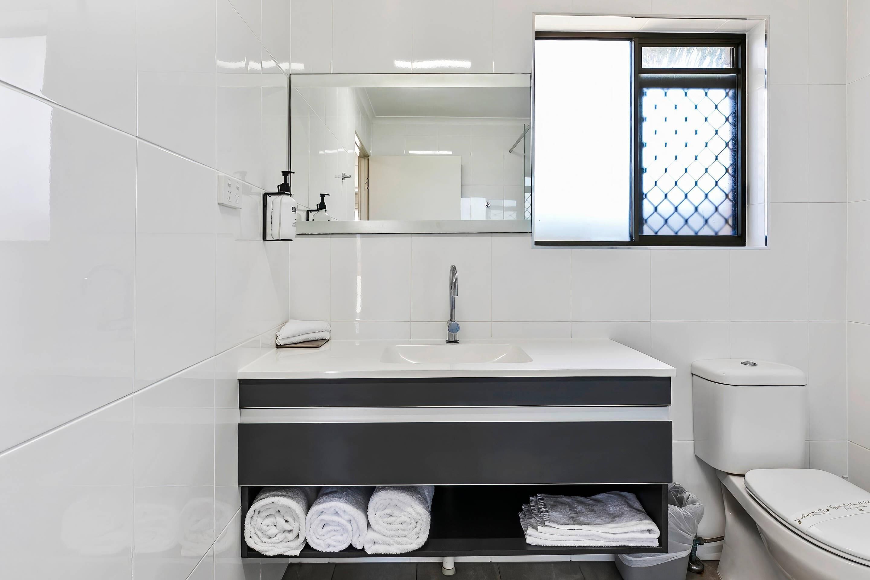 A modern bathroom with a white sink, a mirror, and a toilet. There are towels neatly rolled and stored in a shelf below the sink.