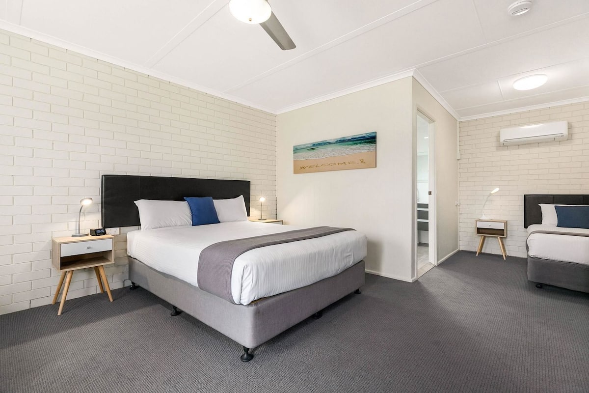 A hotel room with a bed, bedside tables, a nightstand, a wall-mounted air conditioner, and a painting.