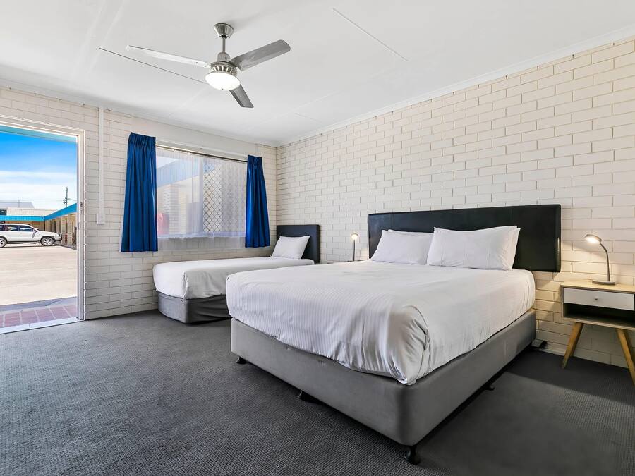 A hotel room with two beds, a ceiling fan, a window with blue curtains, and a view of a parking lot.