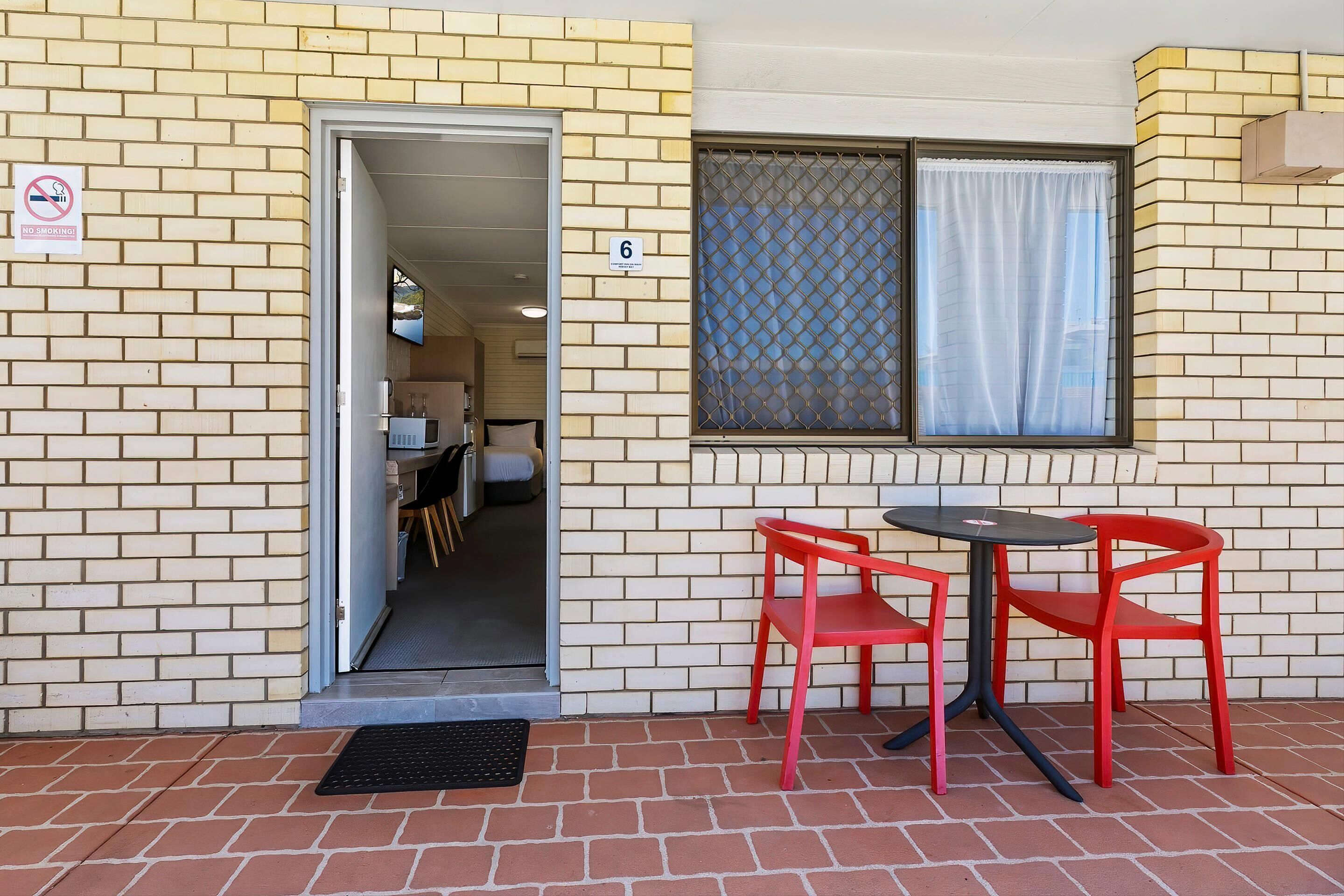 A brick building with a door leading to a room, a window with a curtain, and a table with two red chairs outside.