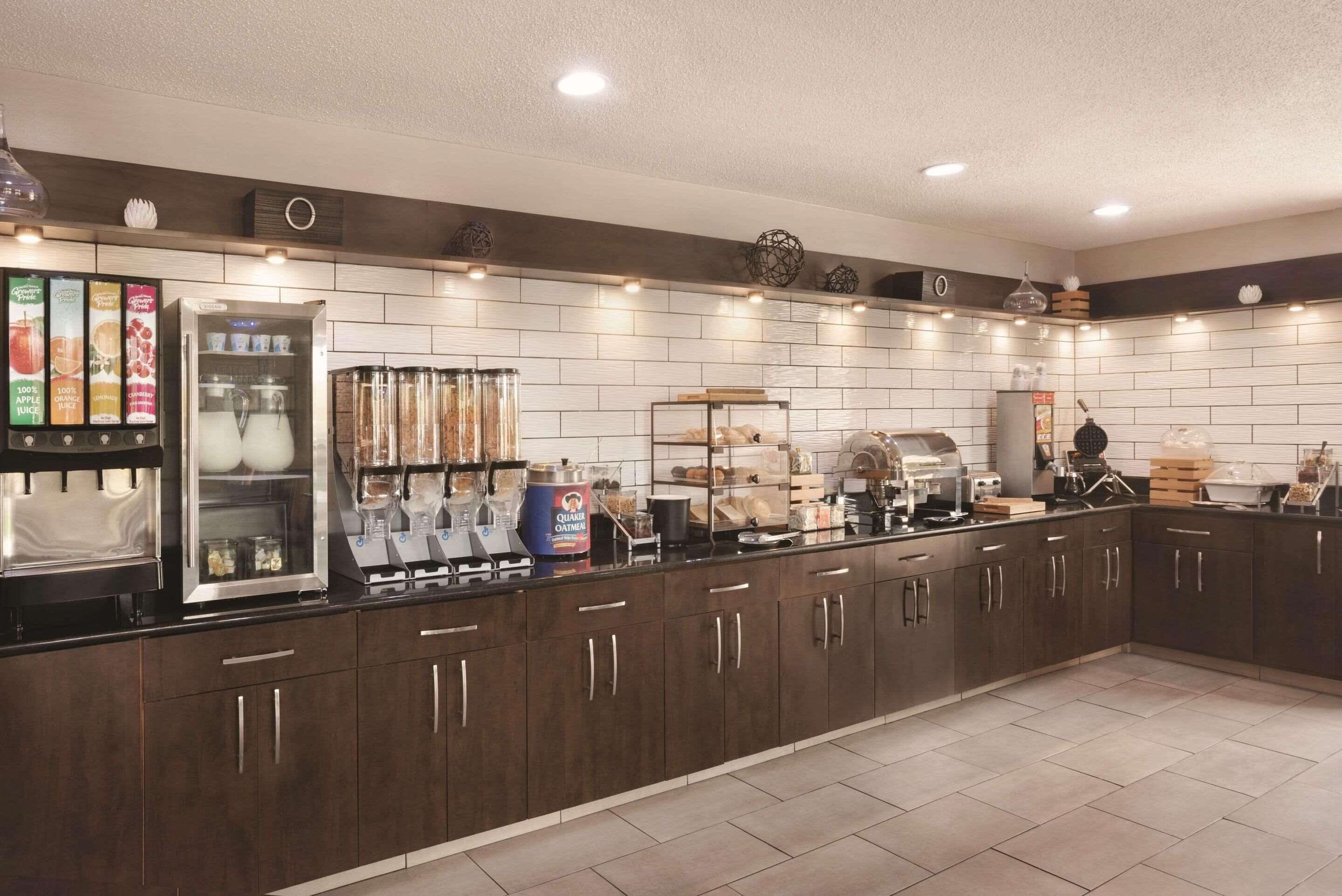 A modern kitchen with a long counter, equipped with a coffee machine, a refrigerator, and various food items.