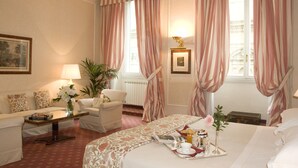 Une chambre d’hôtel comprenant un lit, un coin salon avec un canapé et des fauteuils, une table basse, une lampe et une fenêtre avec des rideaux.