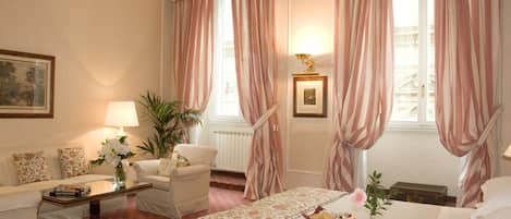 A hotel room with a bed, a sitting area with a sofa and armchairs, a coffee table, a lamp, and a window with curtains.