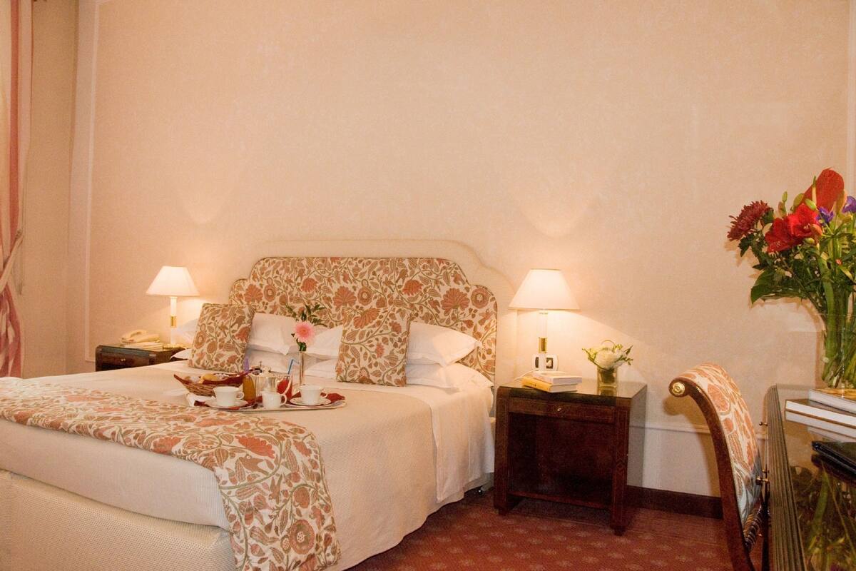 A hotel room with a bed, two bedside tables with lamps, a nightstand, a chair, and a vase with flowers.