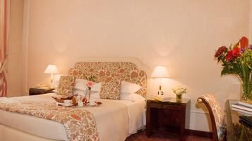 Una camera d'albergo con un letto, due comodini con lampade, una panca e un vaso con fiori.