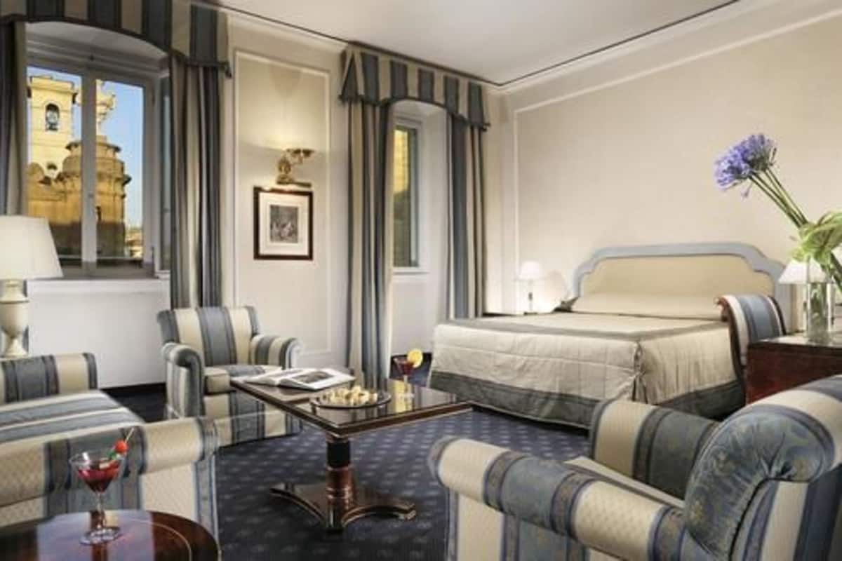 A hotel room with a bed, two armchairs, a small table with a drink, and a view of a historic building.
