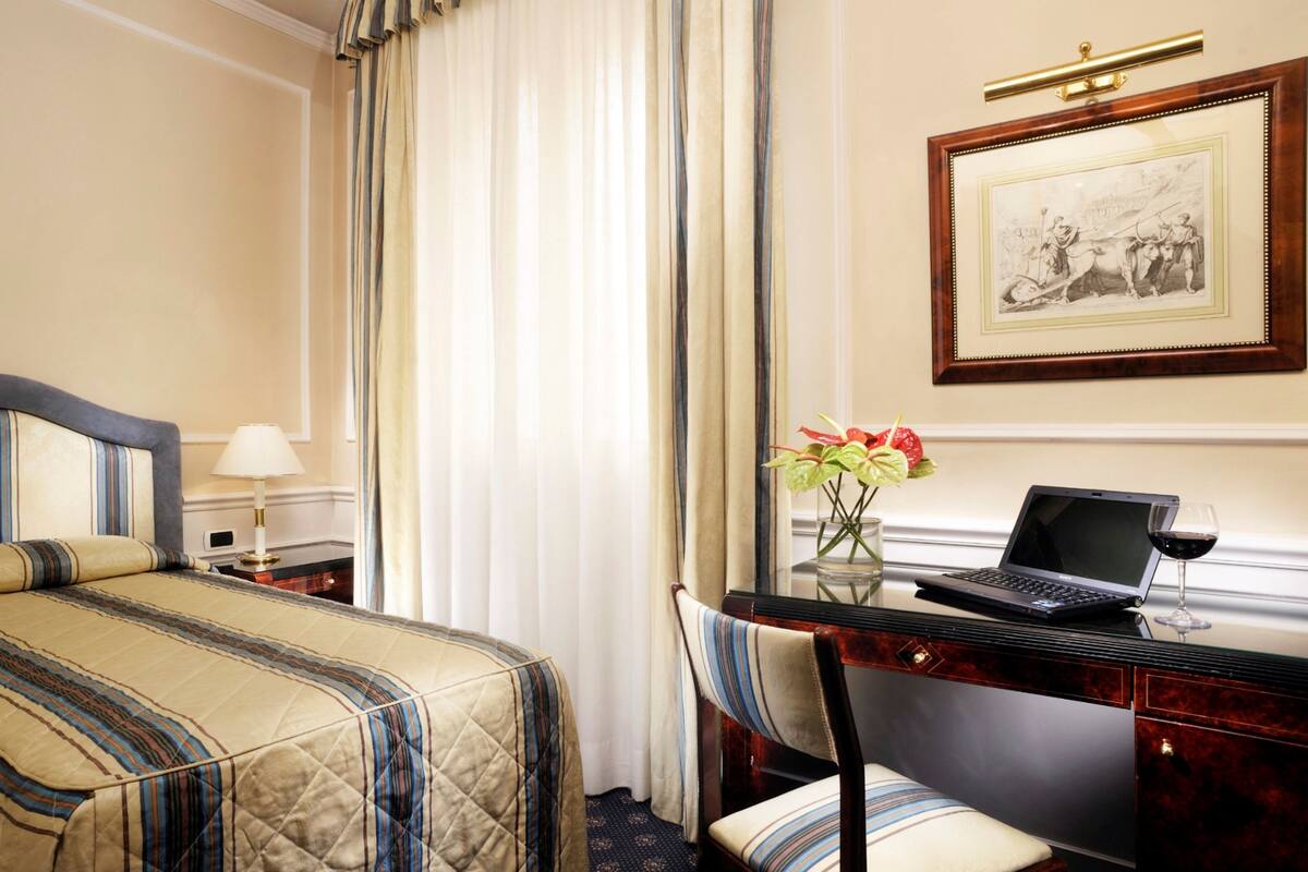 A hotel room with a bed, a desk with a laptop, a vase with flowers, and a framed picture on the wall.