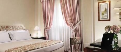 A hotel room with a bed, a chair, a desk with a laptop, a lamp, and a framed picture on the wall.
