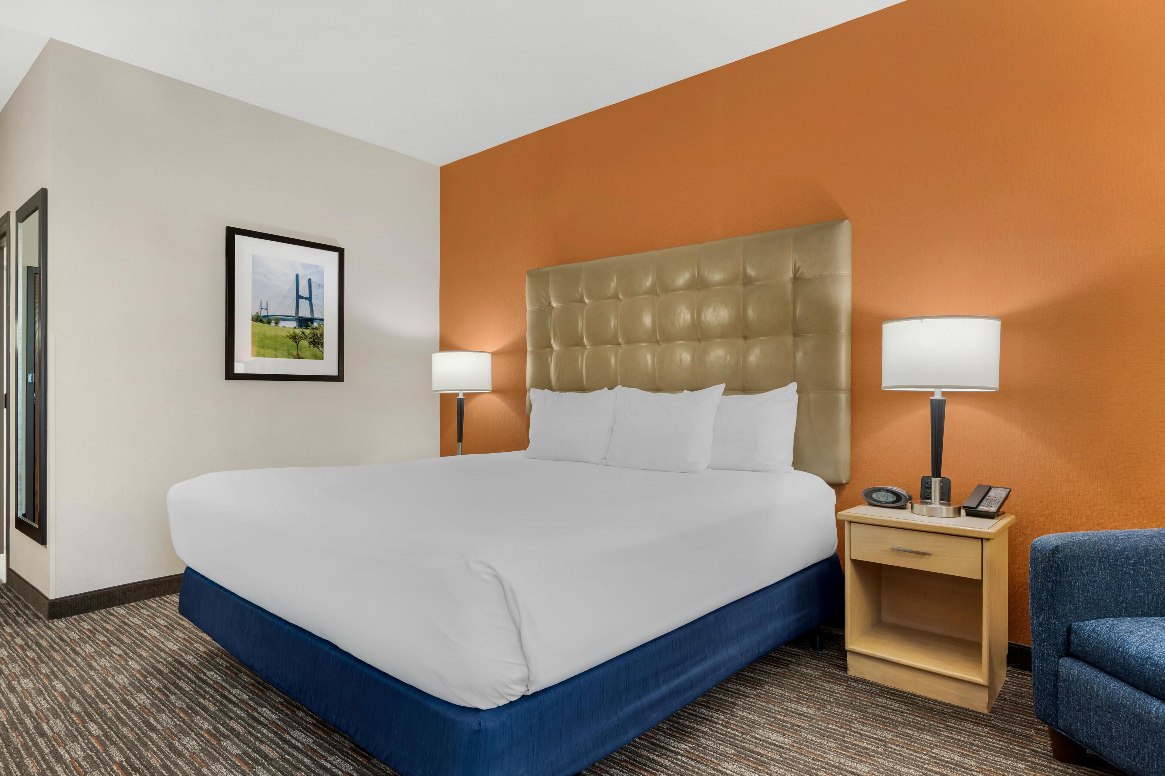 A hotel room with a large bed, two bedside tables with lamps, a framed picture on the wall, and a blue chair.