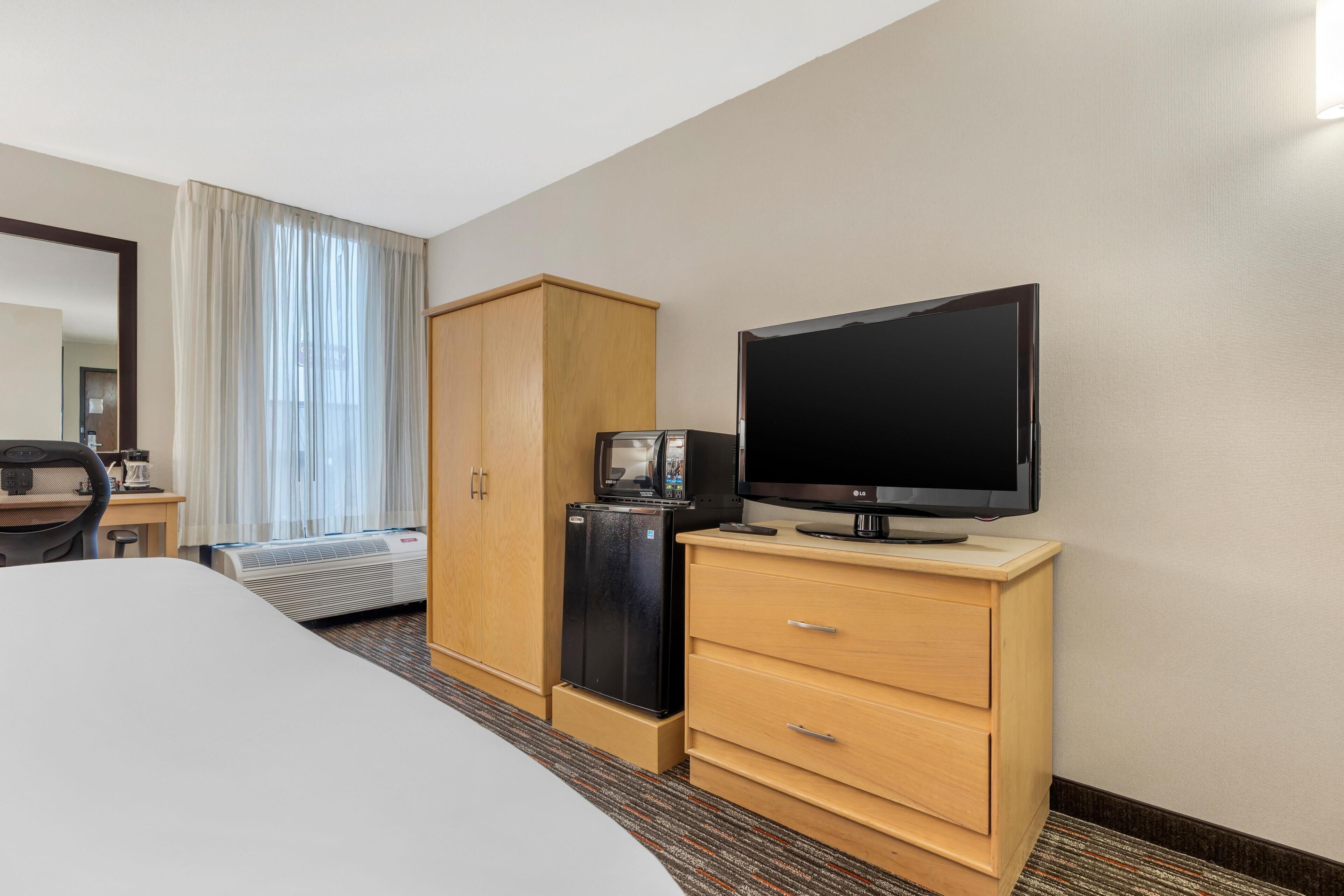 A hotel room with a bed, a television on a wooden stand, a mini-fridge, and a wardrobe.