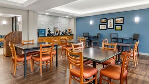 Un'area pranzo moderna con tavoli e sedie in legno, una zona cucina con schienale piastrellato e quadri appesi al muro.