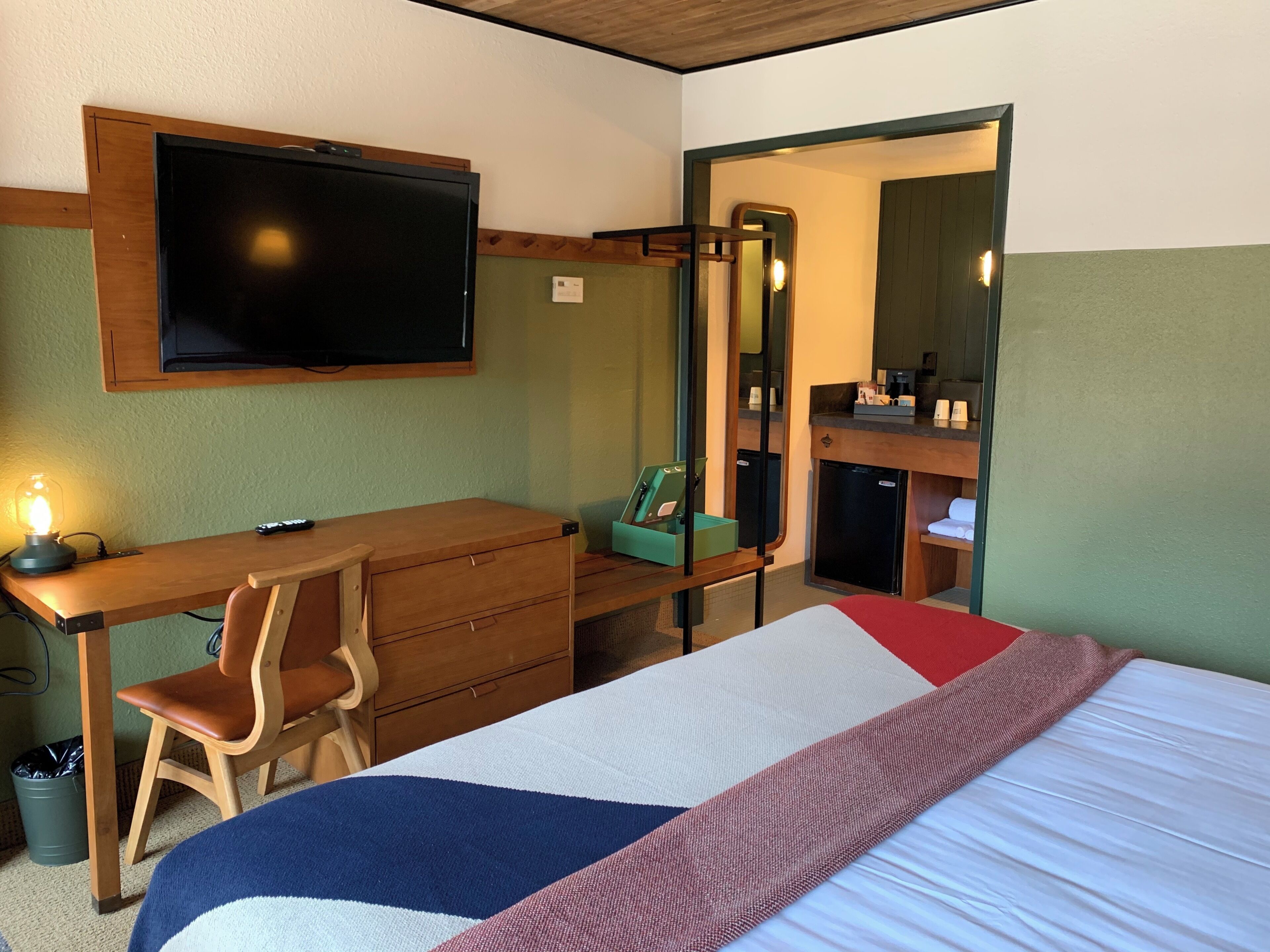 A hotel room with a bed, a desk with a chair, a television, a mirror, and a small kitchenette.