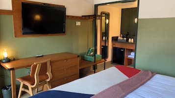 A hotel room with a bed, a desk with a chair, a television, a mirror, and a small kitchenette.
