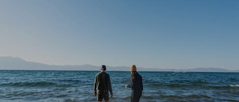 Two people walking on a sandy beach with the ocean and mountains in the background.