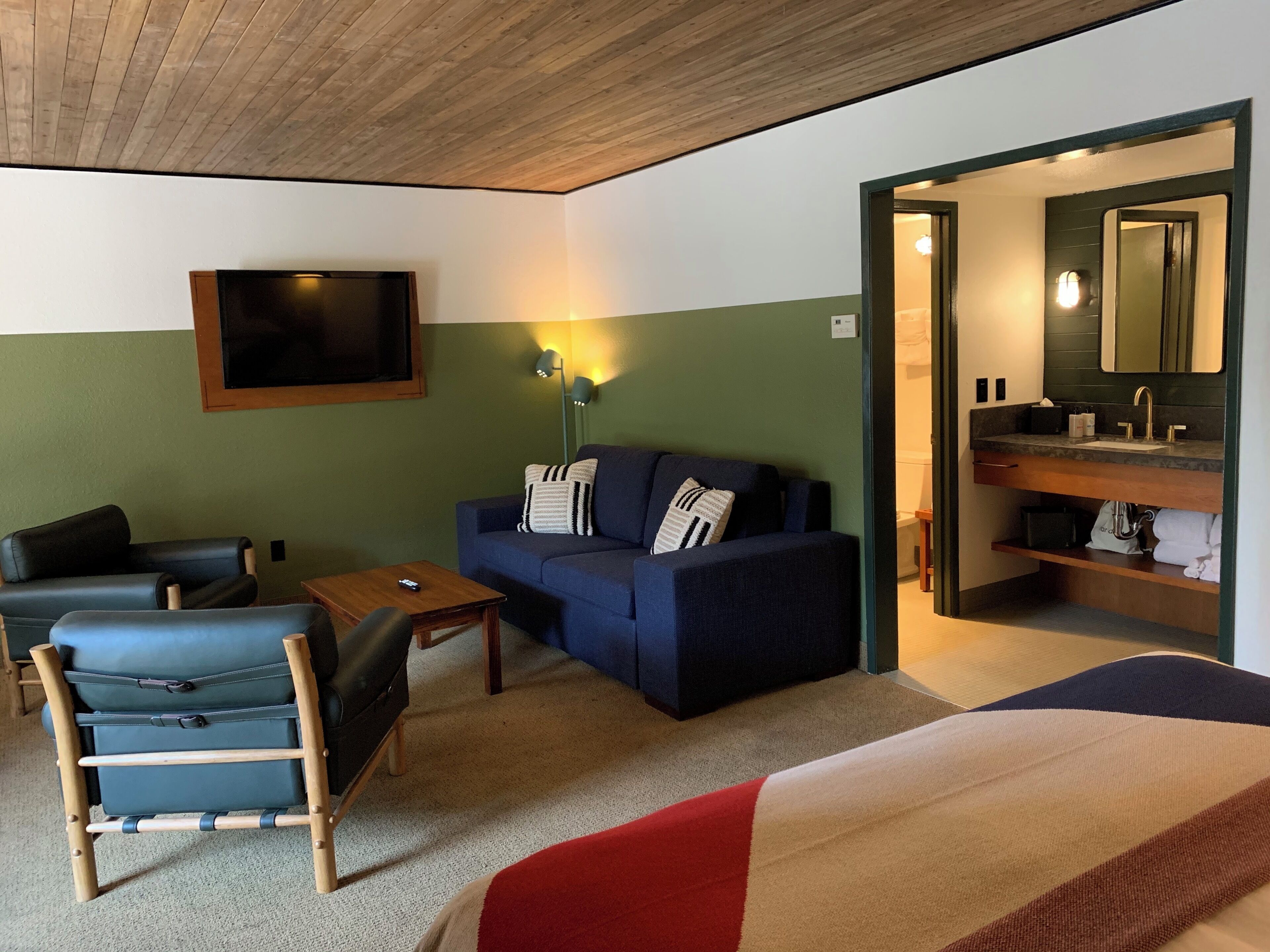 A hotel room with a sofa, a chair, a bed, a TV, a lamp, and a bathroom with a mirror and sink.