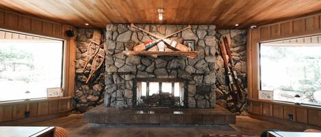 A cozy room with a stone fireplace, wooden ceiling, and large windows.