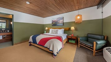Une chambre avec un plafond en bois, un lit recouvert d’une couverture rouge, blanche et bleue, deux lampes de chevet, un fauteuil en cuir vert et un tableau accroché au mur.