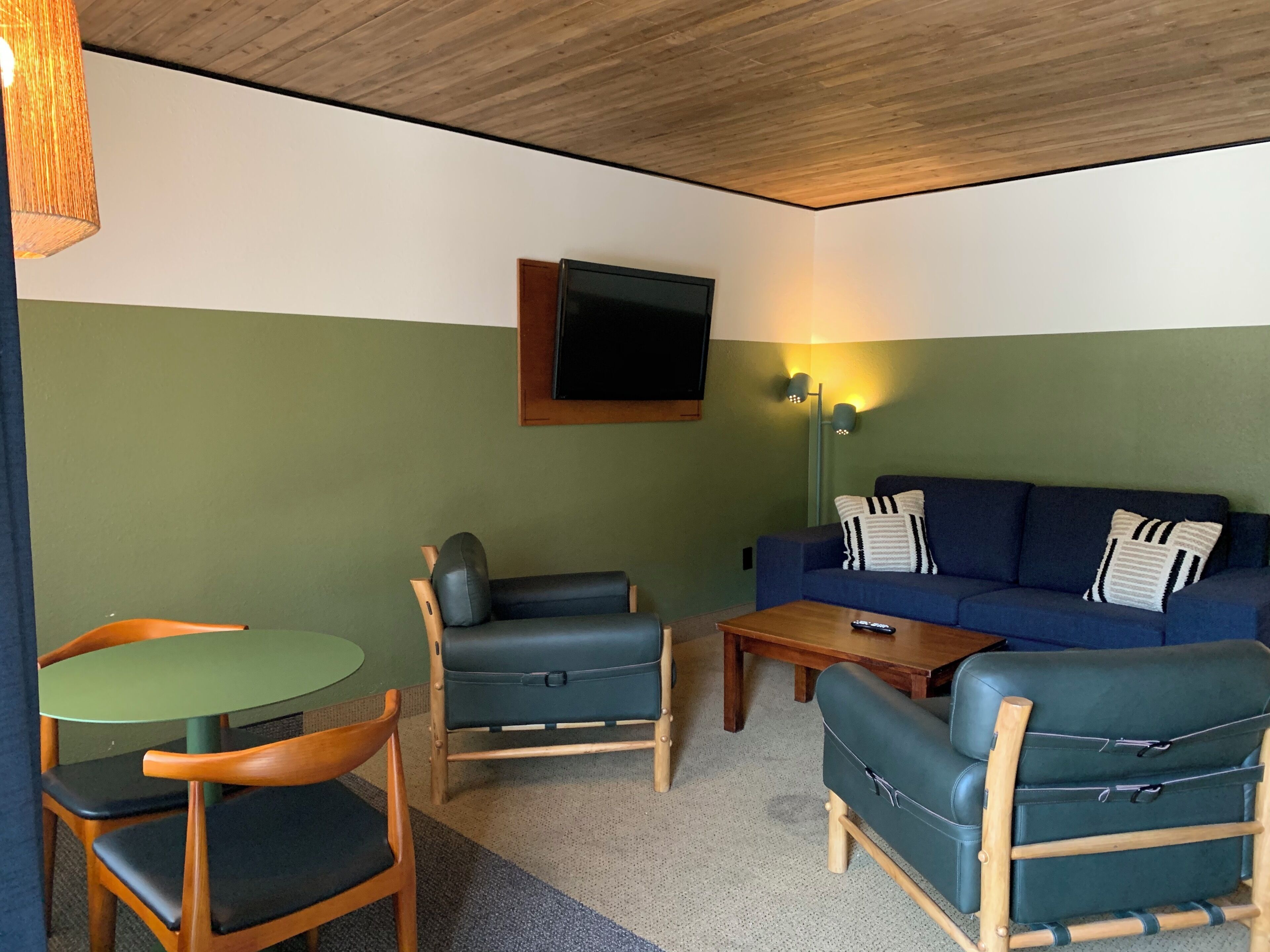 Quarto com parede verde, teto de madeira, sofá azul, duas cadeiras de madeira, uma mesa redonda pequena e uma TV fixada na parede.