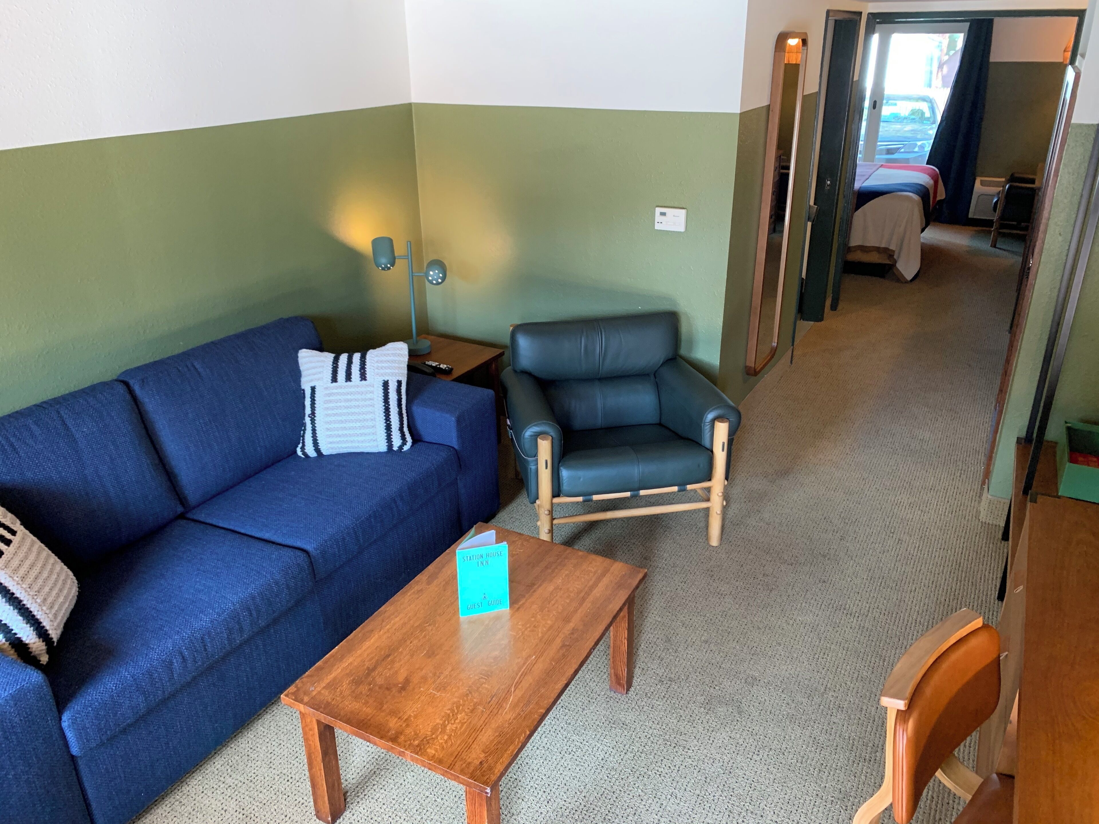 A room with a blue sofa, a wooden table, a black leather chair, and a bed in the background.