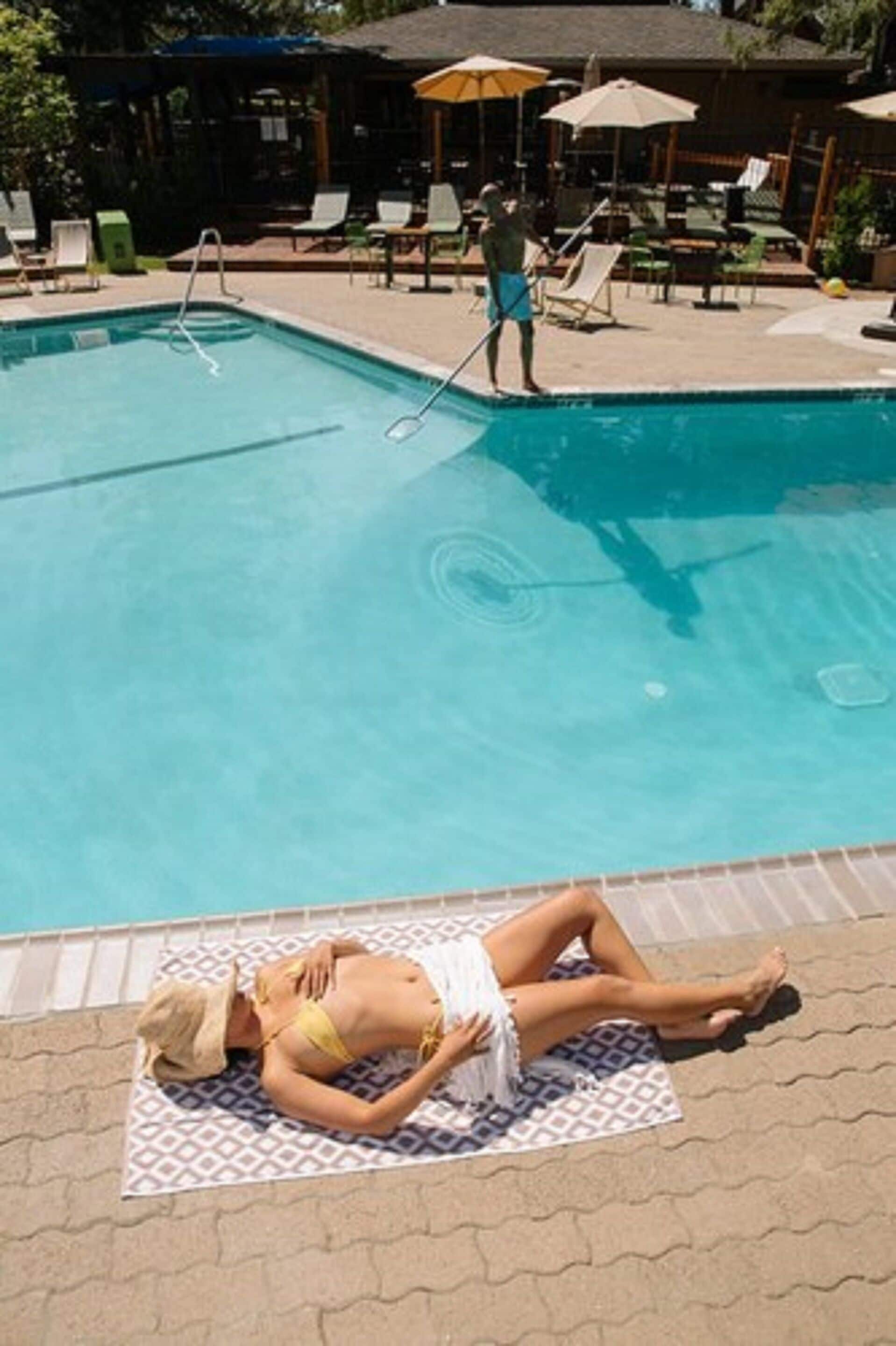A pool area with a person sunbathing on a towel, lounge chairs, and umbrellas.