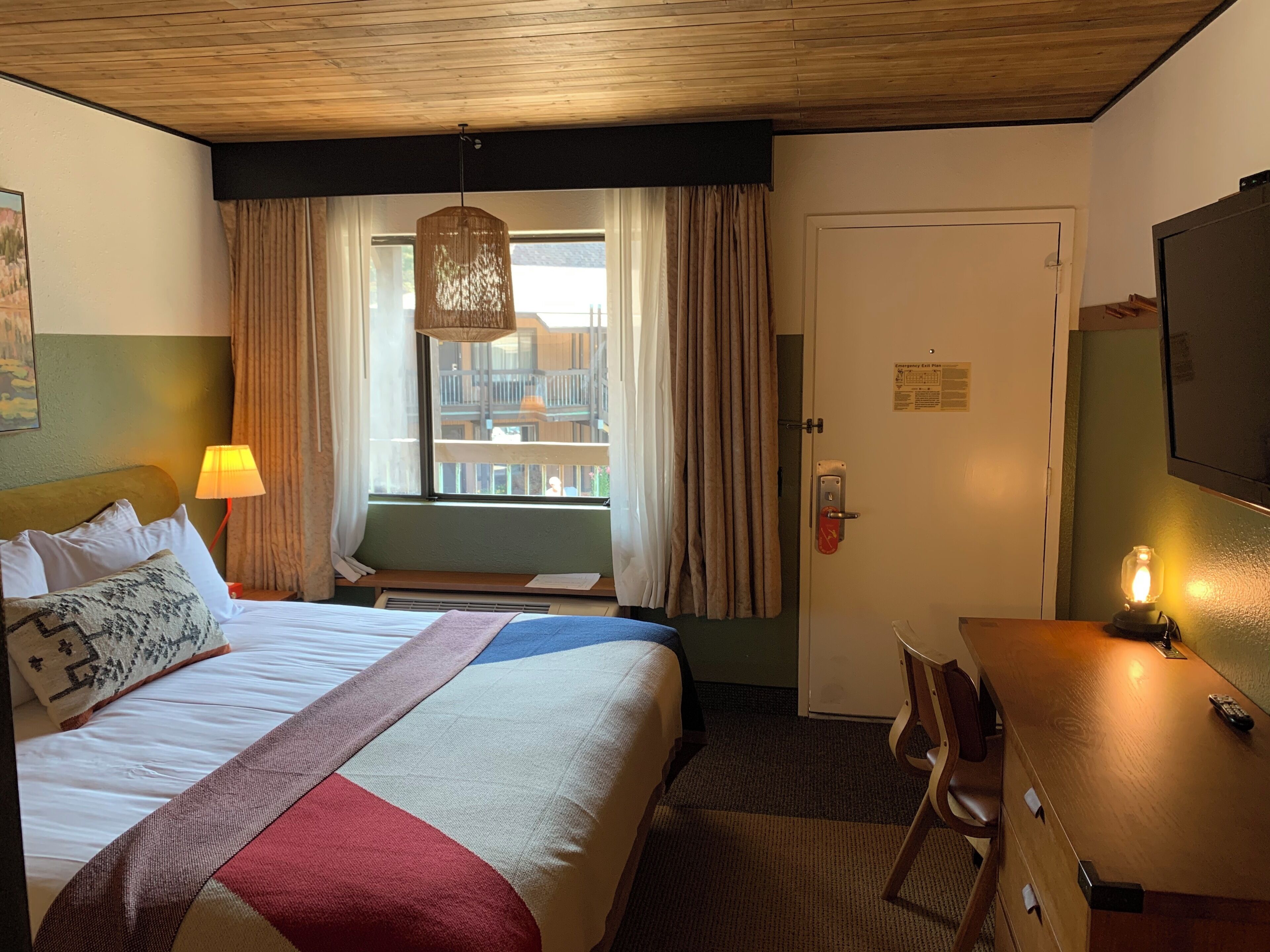 Habitació d'hotel amb un llit, escriptori, cadira, finestra amb cortines, llum penjant i una porta amb el senyal d'emergència.