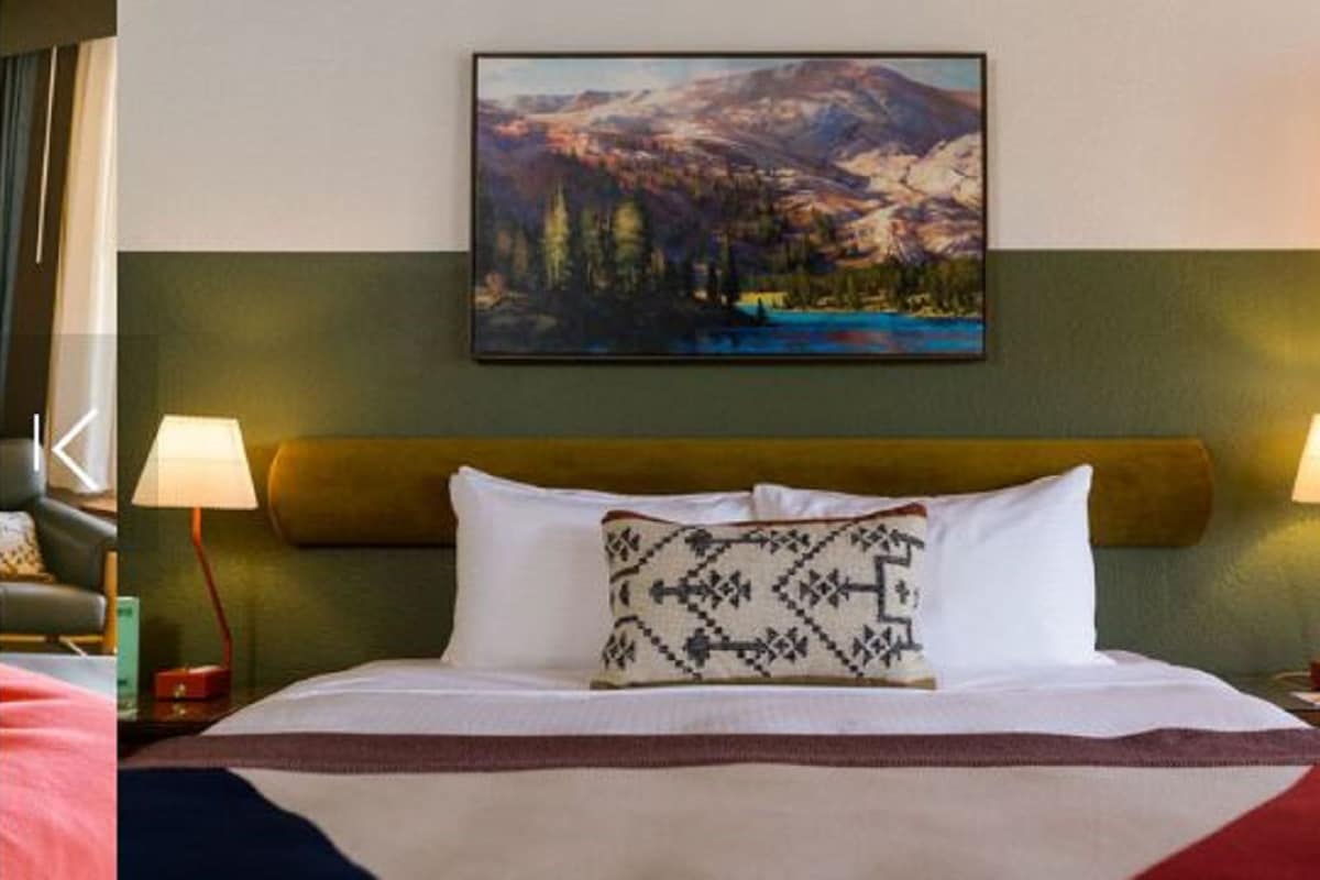 A hotel room with a bed, two bedside lamps, a painting of a mountain landscape, and a patterned throw pillow.