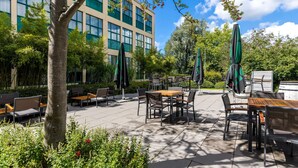An outdoor patio area with tables and chairs, surrounded by greenery and a building with large windows. - NH Amsterdam Noord (Amsterdam)