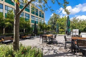 An outdoor patio area with tables and chairs, surrounded by greenery and a building with large windows.
