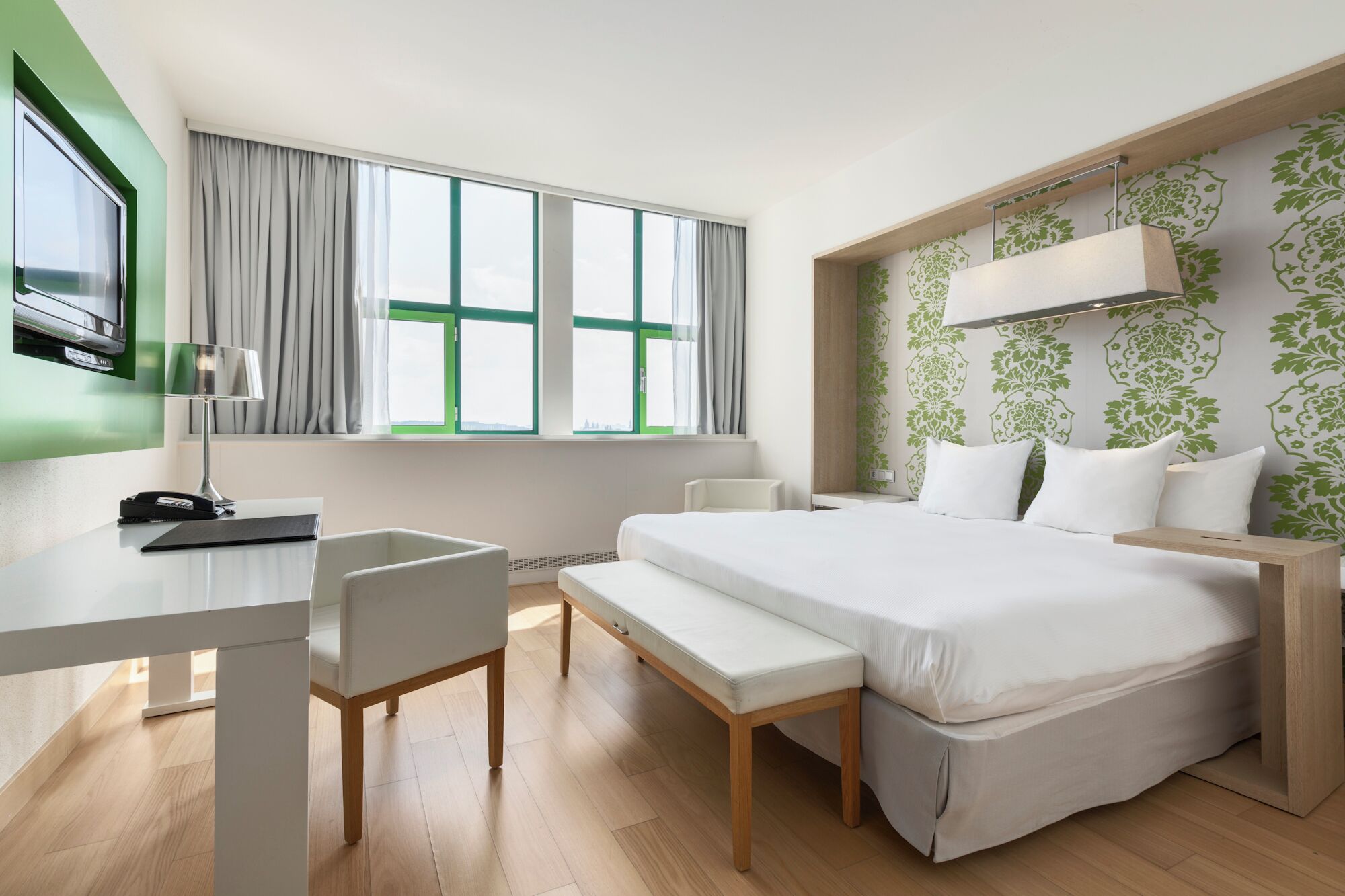 A hotel room with a bed, a desk with a lamp, a chair, and a TV mounted on the wall.