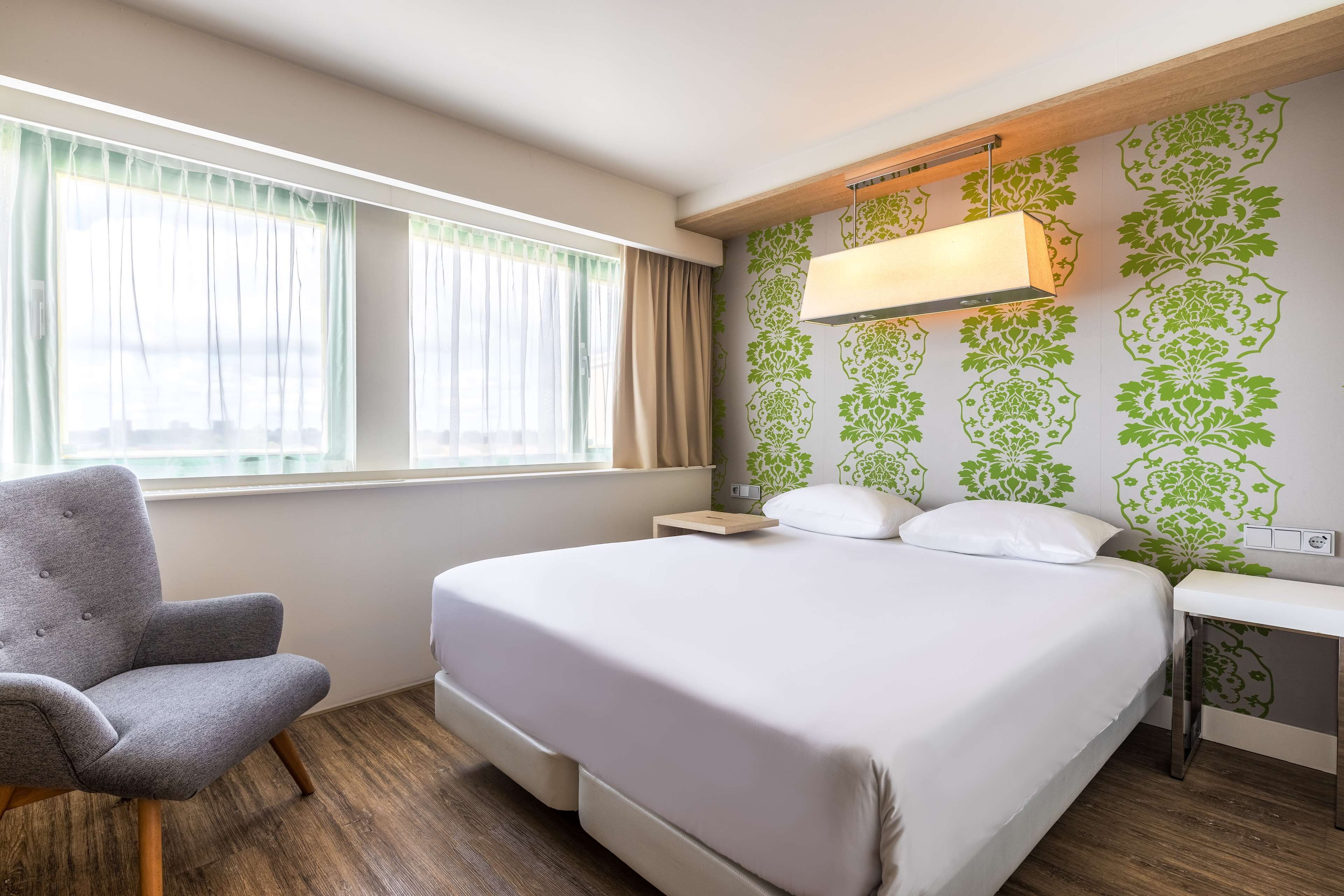 A neatly arranged hotel room with a bed, a chair, a bedside table, and a window with curtains.