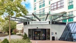The entrance of NH Amsterdam Noord hotel with a glass canopy, a stone wall, and a landscaped area with benches. - NH Amsterdam Noord (Amsterdam)