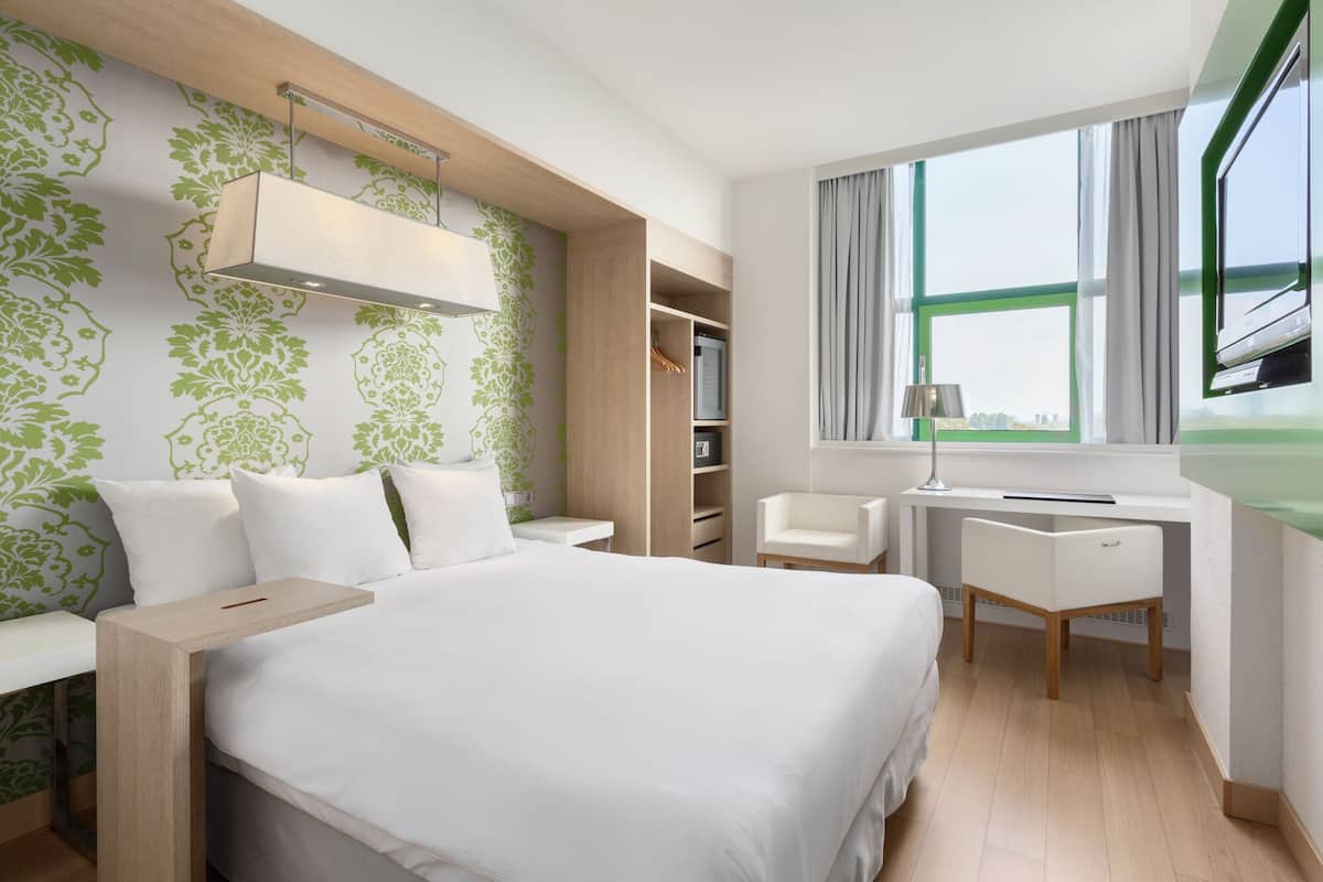 A modern hotel room with a bed, desk, chair, wardrobe, and a window with a view.