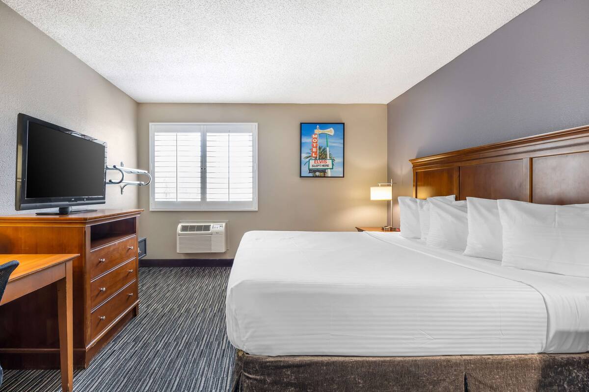 A hotel room with a bed, a desk with a television, a window, and a poster on the wall.