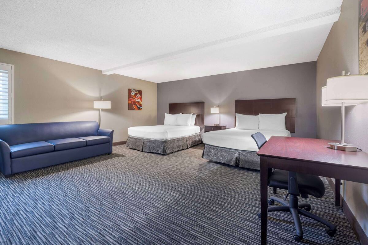A hotel room with a blue sofa, two beds, a desk, and a lamp.